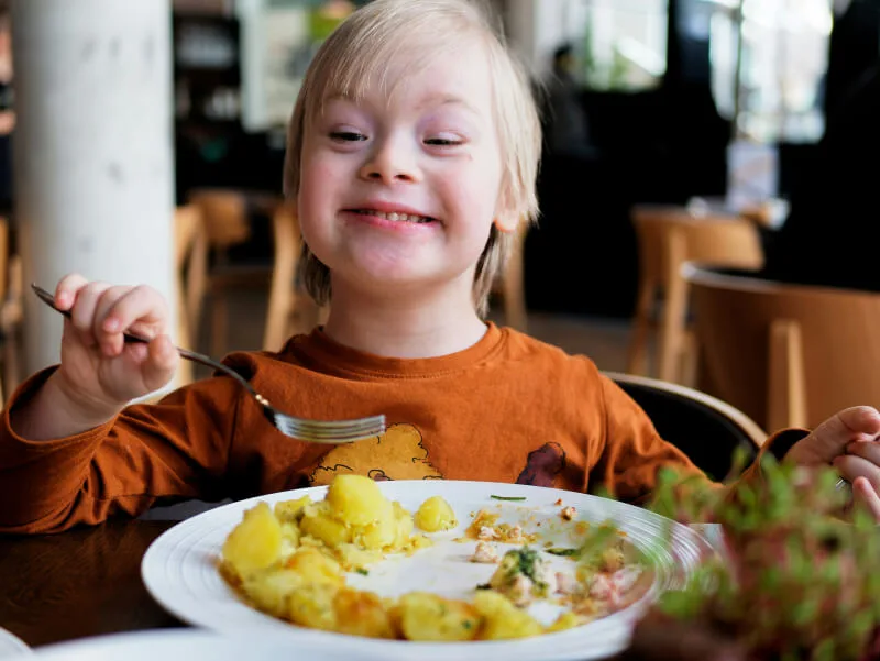 A young child with Down syndrome smiles joyfully while eating a meal of potatoes and chicken at a restaurant, holding a fork and wearing an orange shirt.