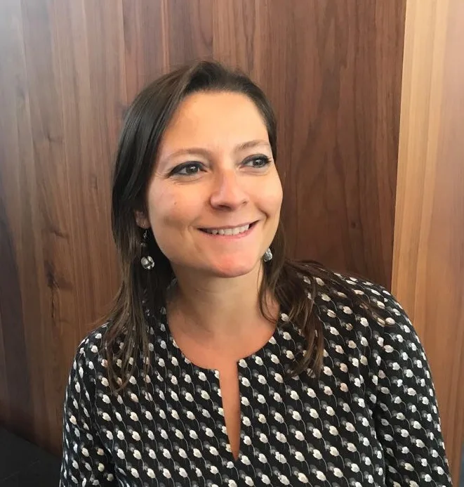 A woman with straight brown hair and dangling earrings smiles while looking slightly to the side. She is wearing a black and white patterned blouse and standing in front of a wooden wall.