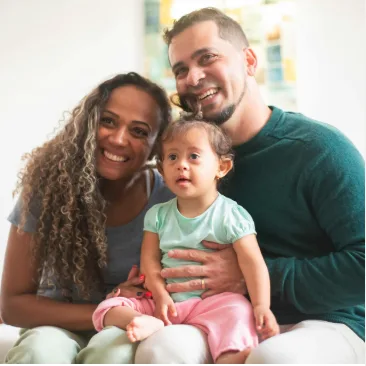 A smiling couple sits closely together indoors, holding a young girl with Down syndrome who is wearing a mint green shirt and pink pants. All three appear happy and relaxed.