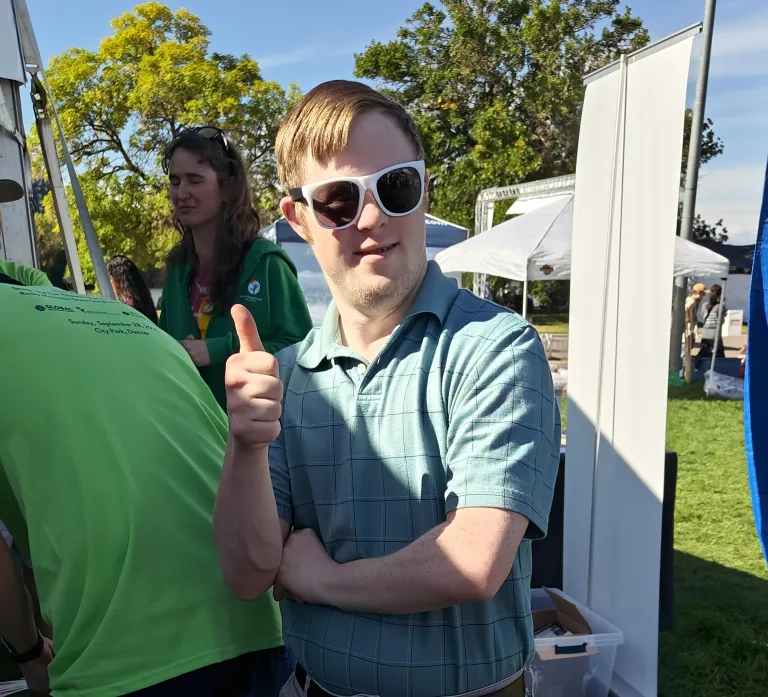 A young man wearing white sunglasses and a blue plaid polo shirt gives a thumbs-up and smiles at an outdoor event, with people and tents in the background.