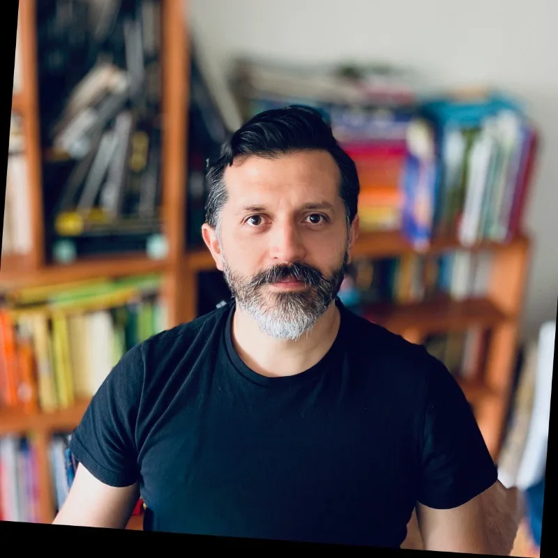 A man with short dark hair and a beard, wearing a black t-shirt, sits in front of shelves filled with colorful books. The background is slightly blurred.