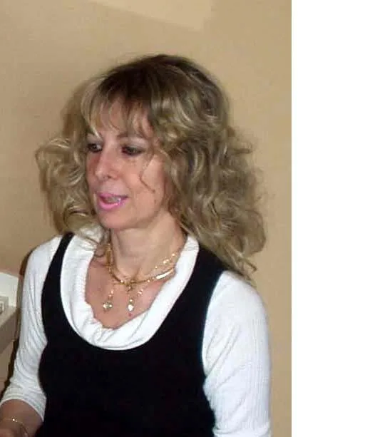 A woman with curly blonde hair, wearing a white long-sleeve top under a black sleeveless dress and a gold necklace, is smiling slightly while looking down. She is indoors against a beige wall.